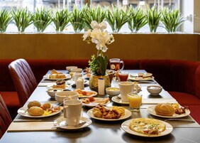 Een eettafel gedekt met diverse ontbijtartikelen, zoals eieren, toast en fruit, in een ruimte met rode zitplaatsen en planten op de achtergrond.