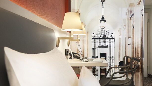 A hotel room with a bed, a desk with a lamp, a chair, and a view of a decorative gate and arches. - Best Western Hotel Leipzig City Center (Leipzig)