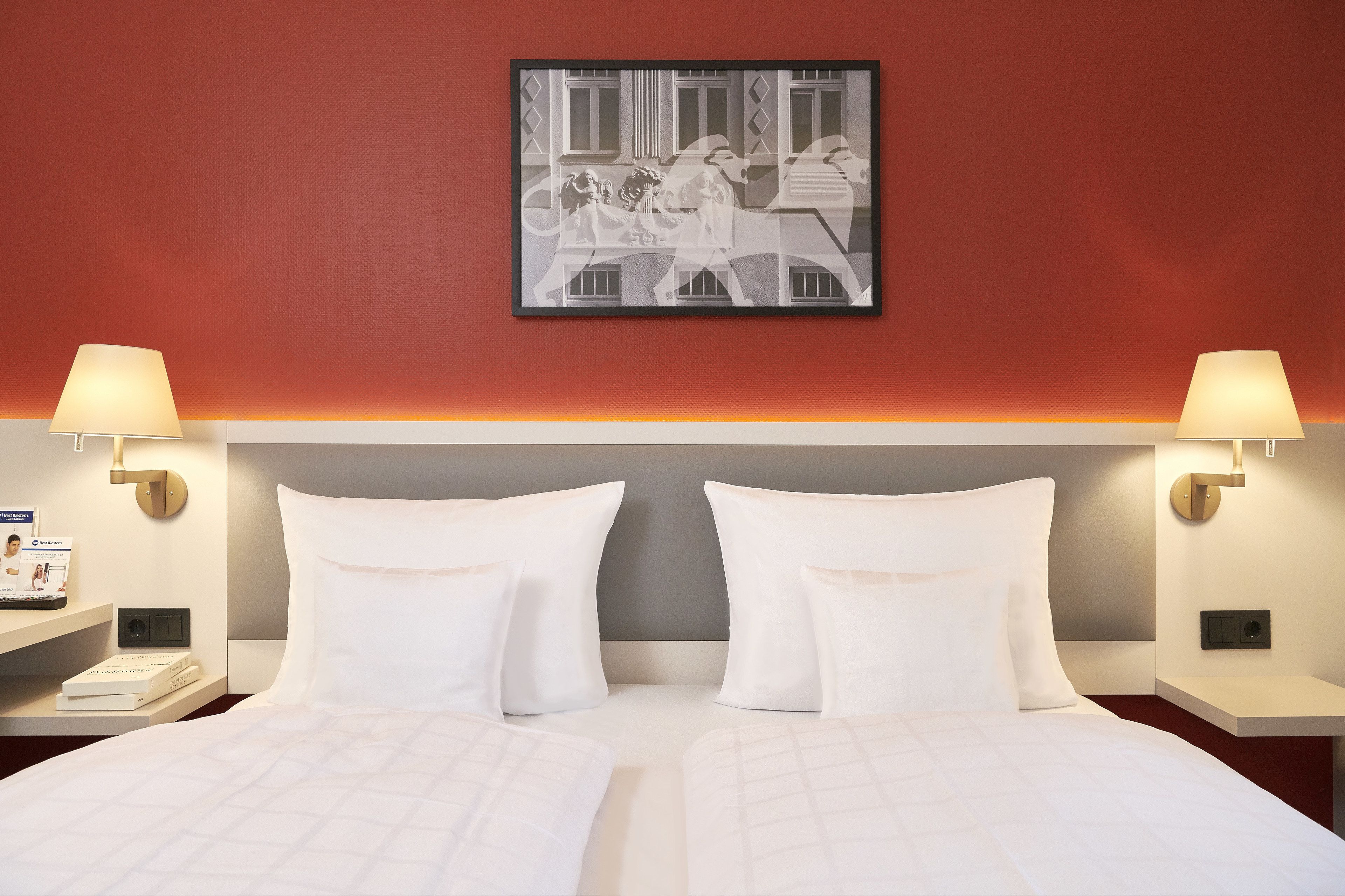 Habitación de hotel con una cama, dos mesitas de noche con lámparas, una obra de arte en la pared y una mesita con un teléfono y una revista.