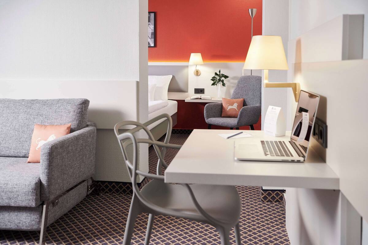 Habitación de hotel moderna con un sofá gris, un escritorio blanco con una computadora portátil, un sillón gris y una pared de acento roja.