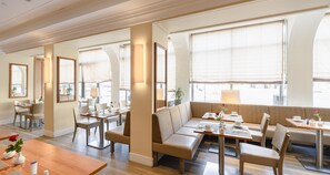 Un comedor moderno con mesas, sillas y un sofá. Amplias ventanas proporcionan luz natural.