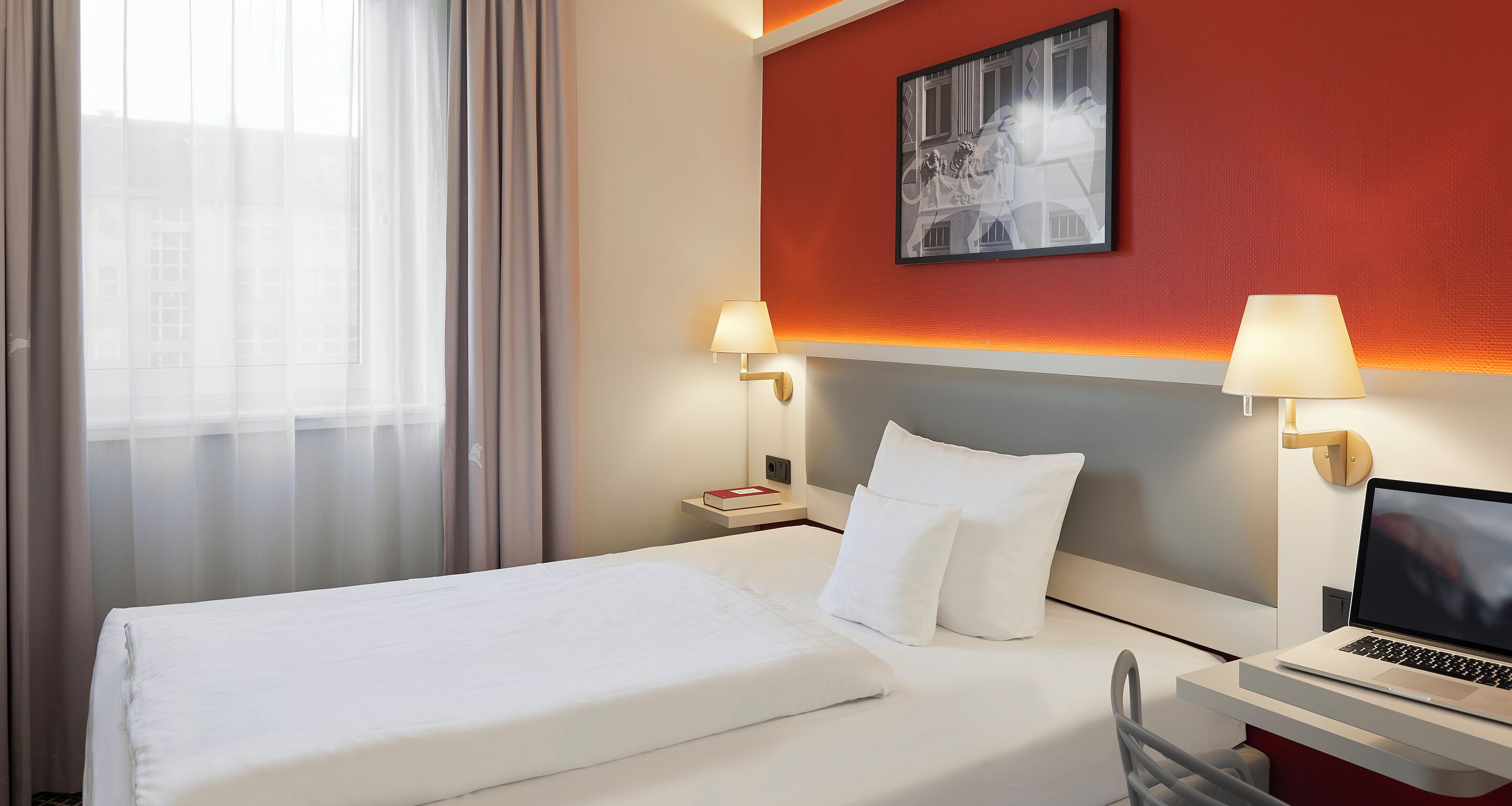 Habitaci ón de hotel con una cama, dos lámparas de noche, un ordenador portátil sobre una mesita y un cuadro en la pared.