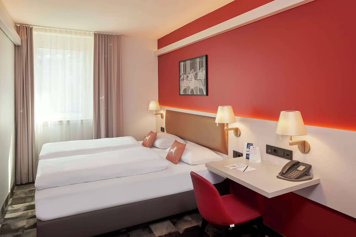 Una habitación de hotel con una cama, dos lámparas de noche, un escritorio con teléfono y una silla roja.