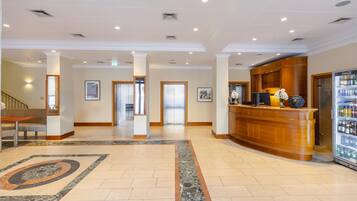 Un hall de hotel con una mesa de recepción, un refrigerador lleno de bebidas y una escalera que sube a otro piso.