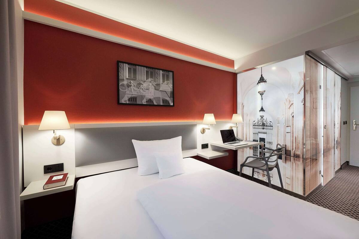 Habitación de hotel con una cama grande, dos mesitas de noche con lámparas, un escritorio con una computadora portátil, un cuadro en la pared y un armario con espejo.