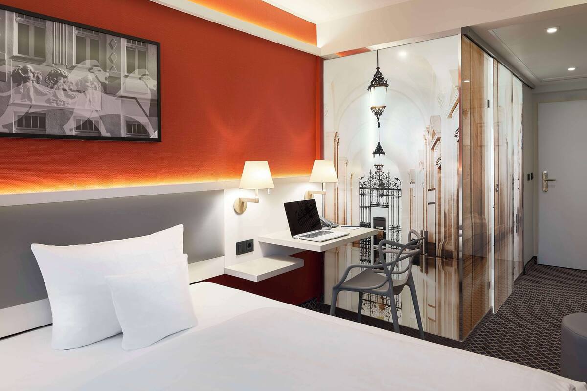 Habitación de hotel con una cama, un escritorio con una computadora portátil, dos lámparas y un cuadro grande en la pared.