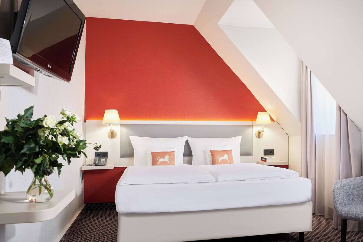 Habitación de hotel con cama, sillón, televisor, escritorio con teléfono y un jarrón con flores.