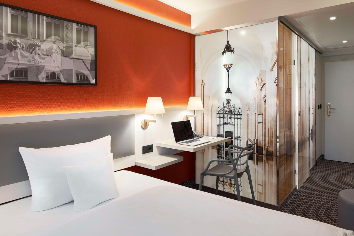 Ein Hotelzimmer mit einem Bett, einem Schreibtisch mit einem Laptop, zwei Lampen und einem großen Spiegel.