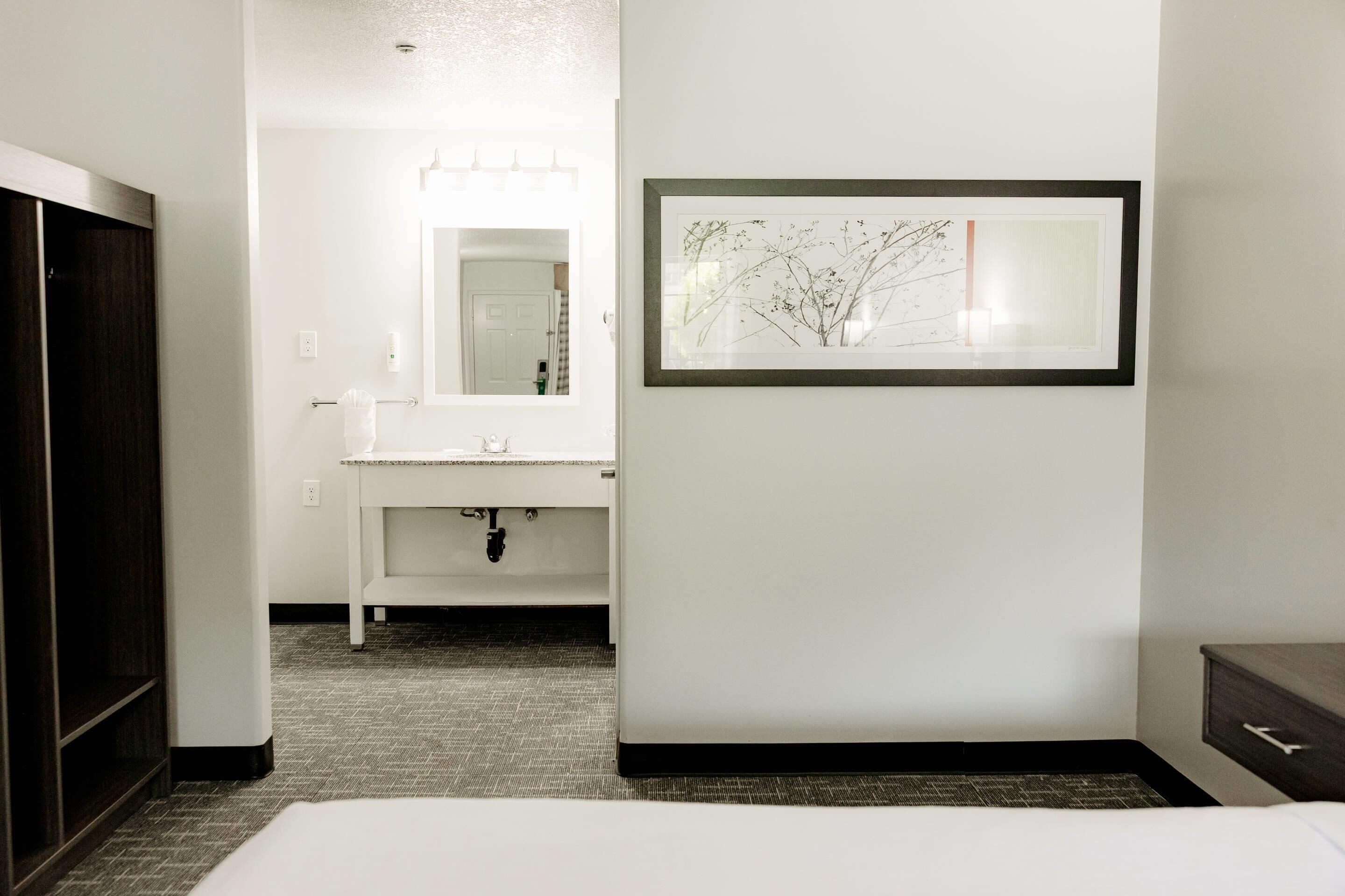 Ein Hotelzimmer mit Bett, Badezimmer mit Waschbecken und Spiegel, sowie ein botanisches Bild in einem Rahmen an der Wand.