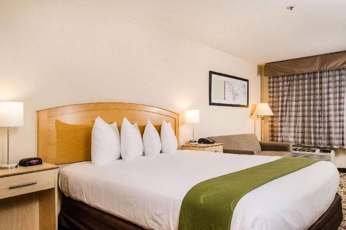 A hotel room with a large bed, two bedside tables, a chair, a desk, a lamp, a phone, and a window with curtains.