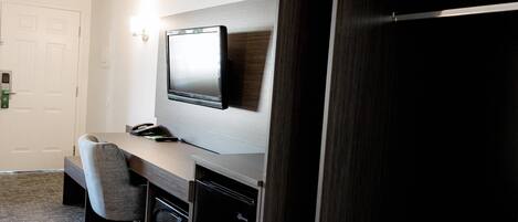 A hotel room with a flat-screen TV mounted on the wall, a desk with a chair, a microwave, and a refrigerator.
