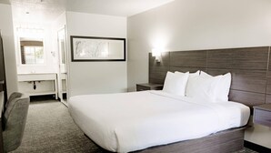 A hotel room with a large bed, a bedside table, a nightstand, a chair, a bathroom with a sink and mirror, and a framed artwork on the wall. - Quality Inn & Suites Lathrop - South Stockton (Lathrop)