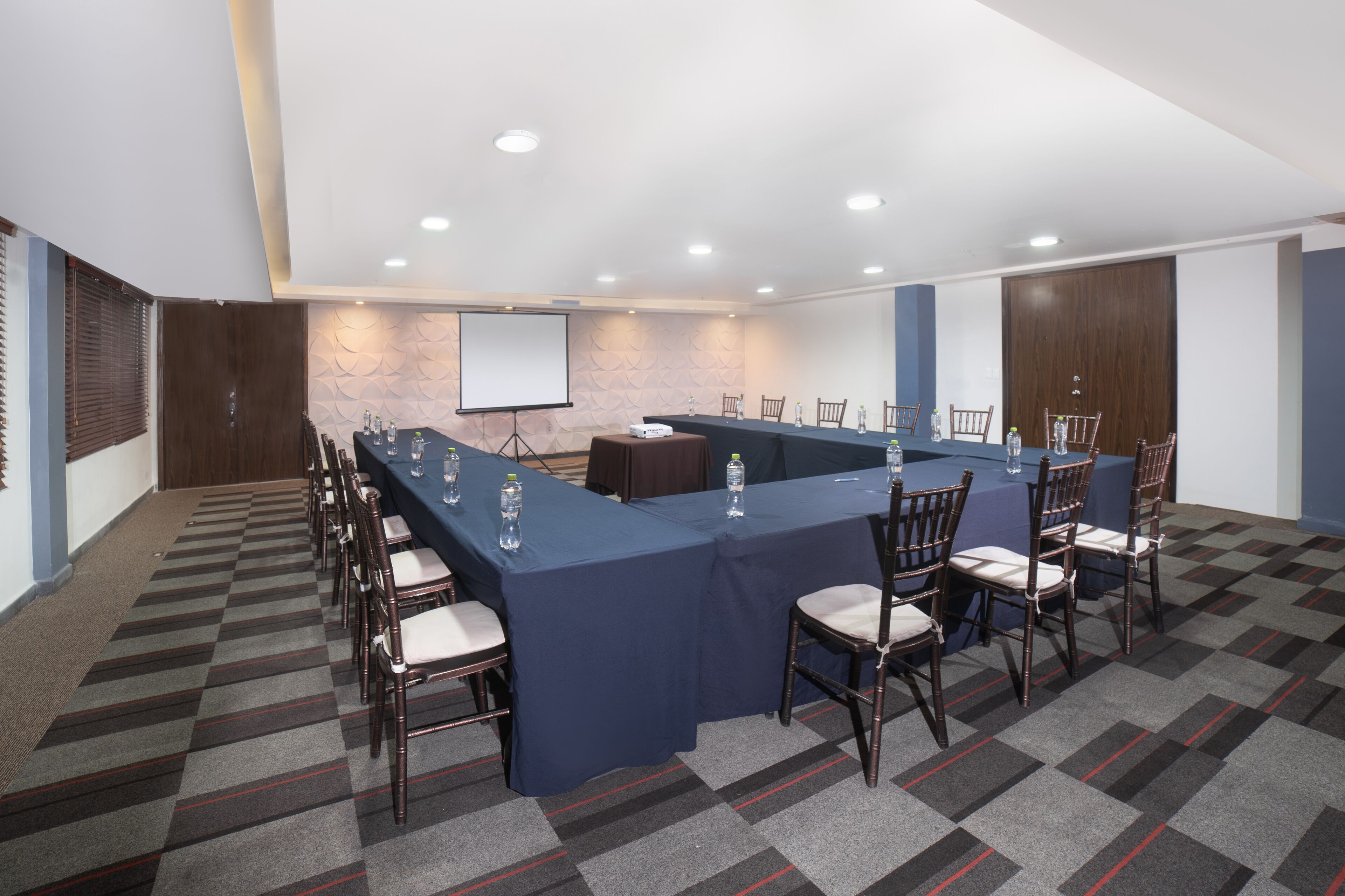 Un salón de conferencias con una larga mesa, sillas, una pantalla de proyector y suelo alfombrado.
