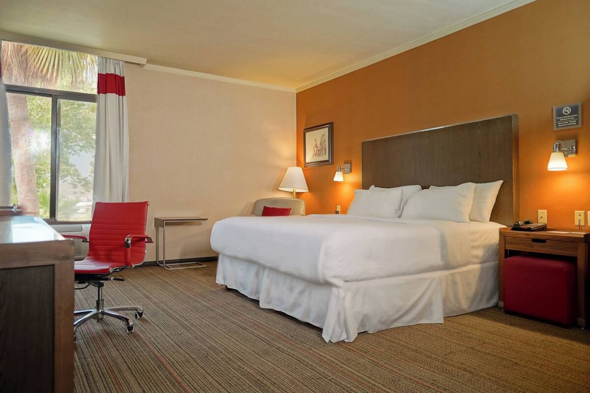 Habitación de hotel con una cama grande, un escritorio con silla, una lámpara y un ventanal con cortinas.