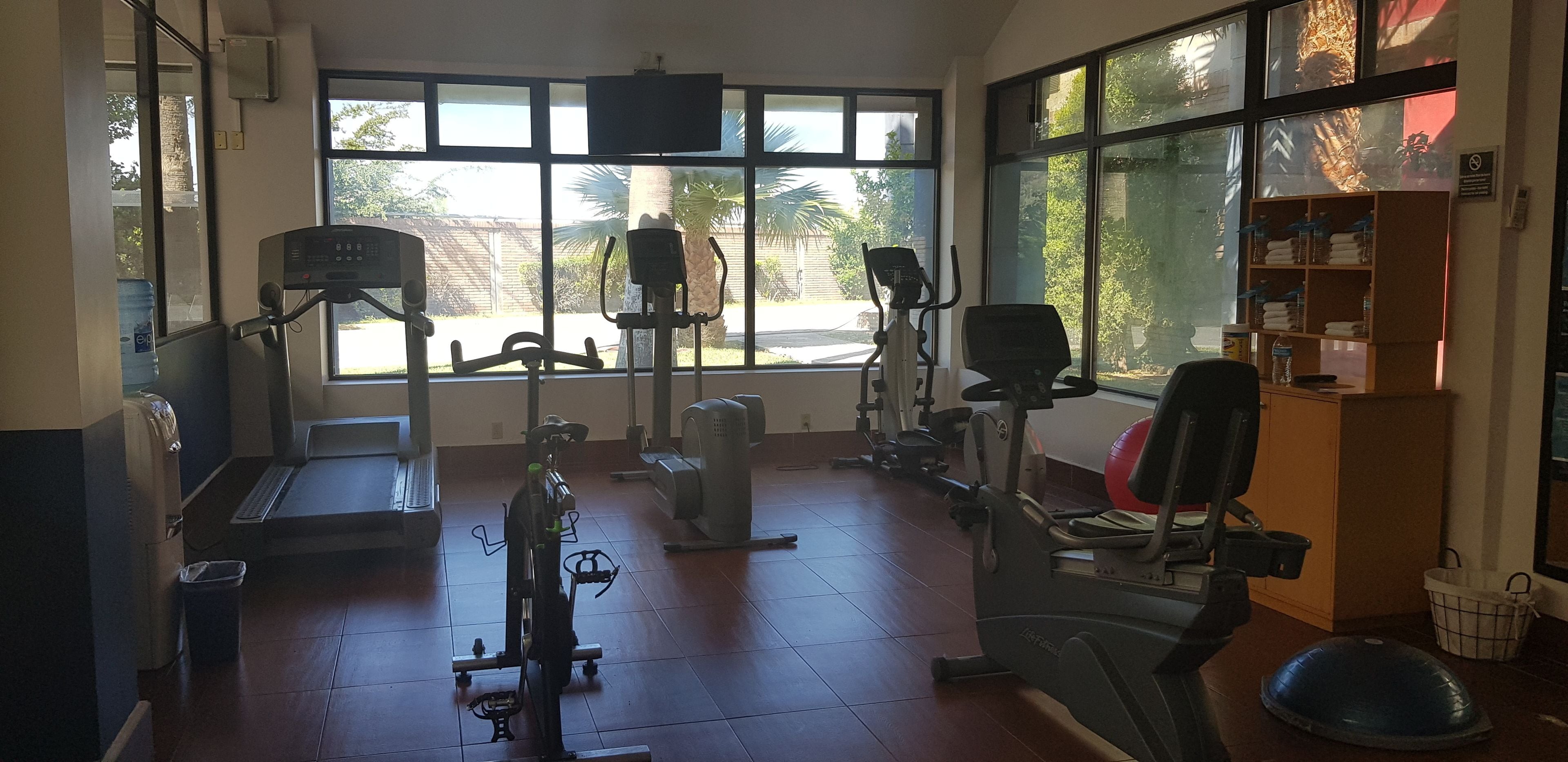 A gym with various exercise equipment, large windows, and a wooden shelf with towels.