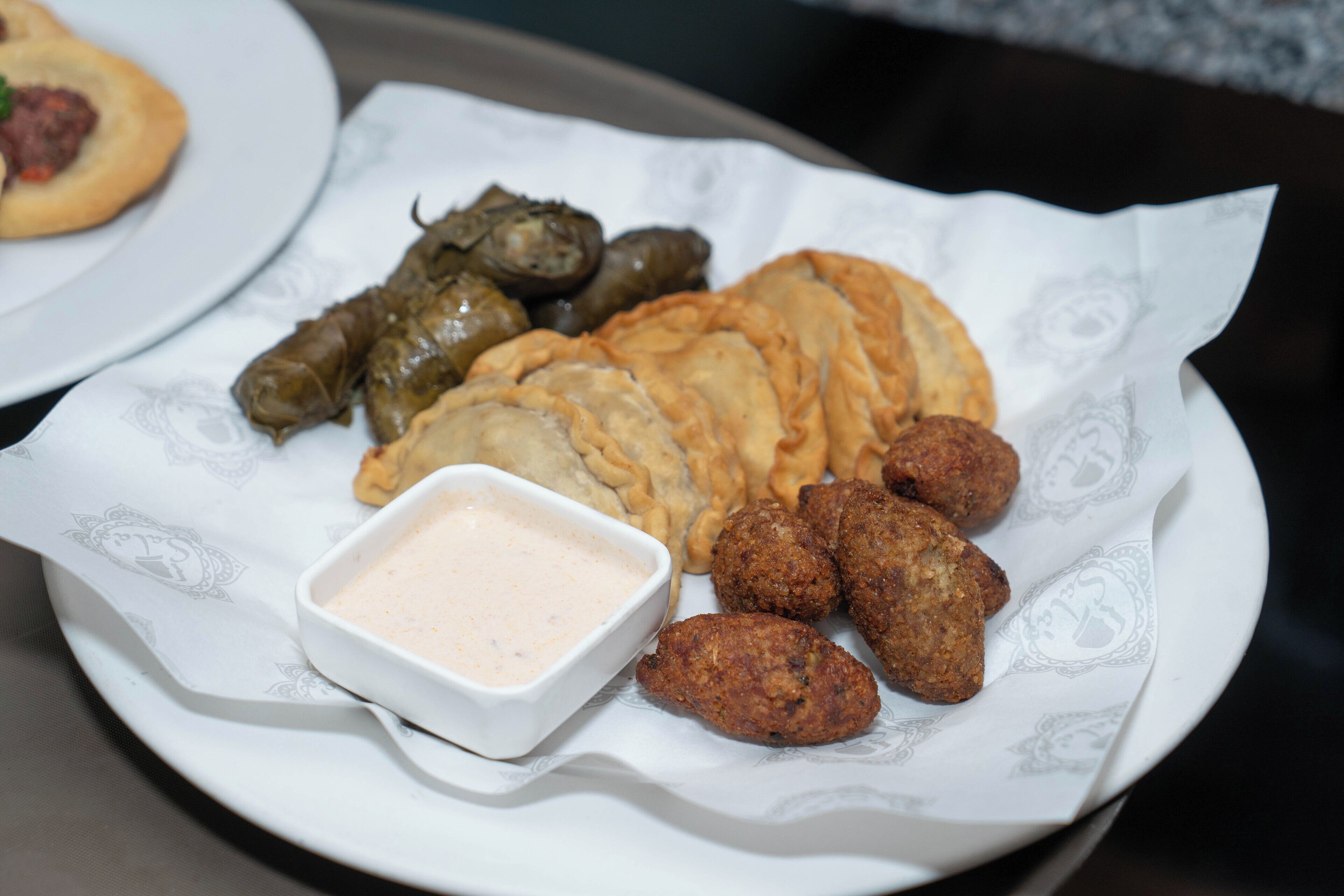 Une assiette de mets comprenant des feuilles de vigne farcies, des pâtisseries frites et des boulettes de viande accompagnées d’une sauce pour tremper.