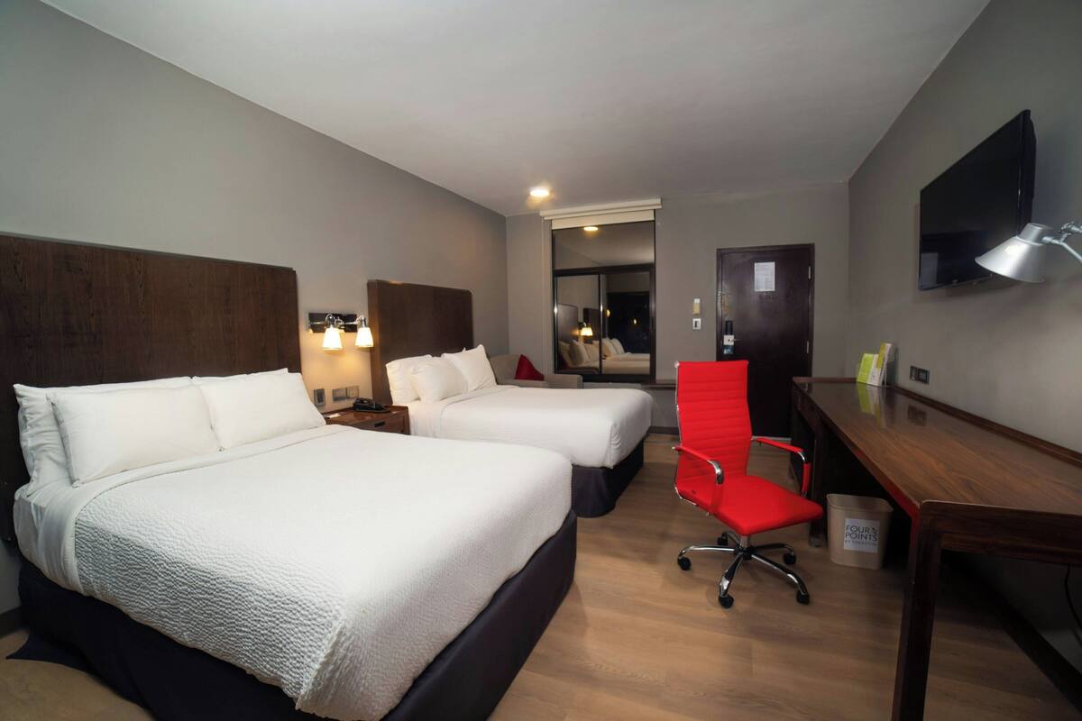 Habitación de hotel con dos camas, un escritorio, una silla, un televisor y un baño visible a través de una puerta.