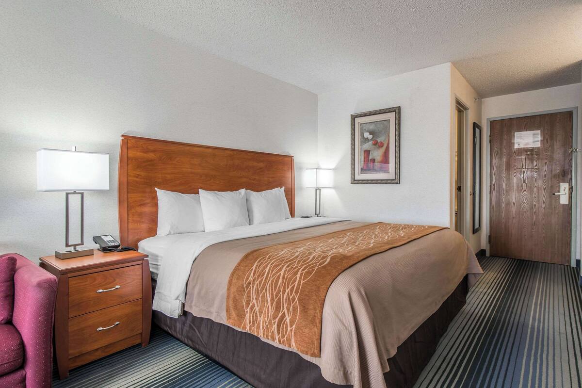 A hotel room with a large bed, a wooden nightstand with a phone, a desk lamp, a chair, and a framed picture on the wall.