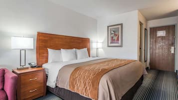Een hotelkamer met een groot bed, een houten nachtkastje met een telefoon, een bureaulamp, een stoel en een ingelijst schilderij aan de muur.