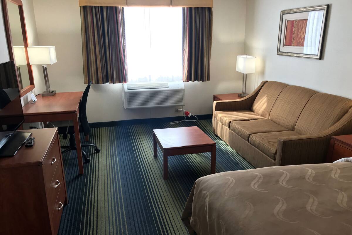 A hotel room with a bed, a sofa, a desk with a computer, a chair, and a window with curtains.