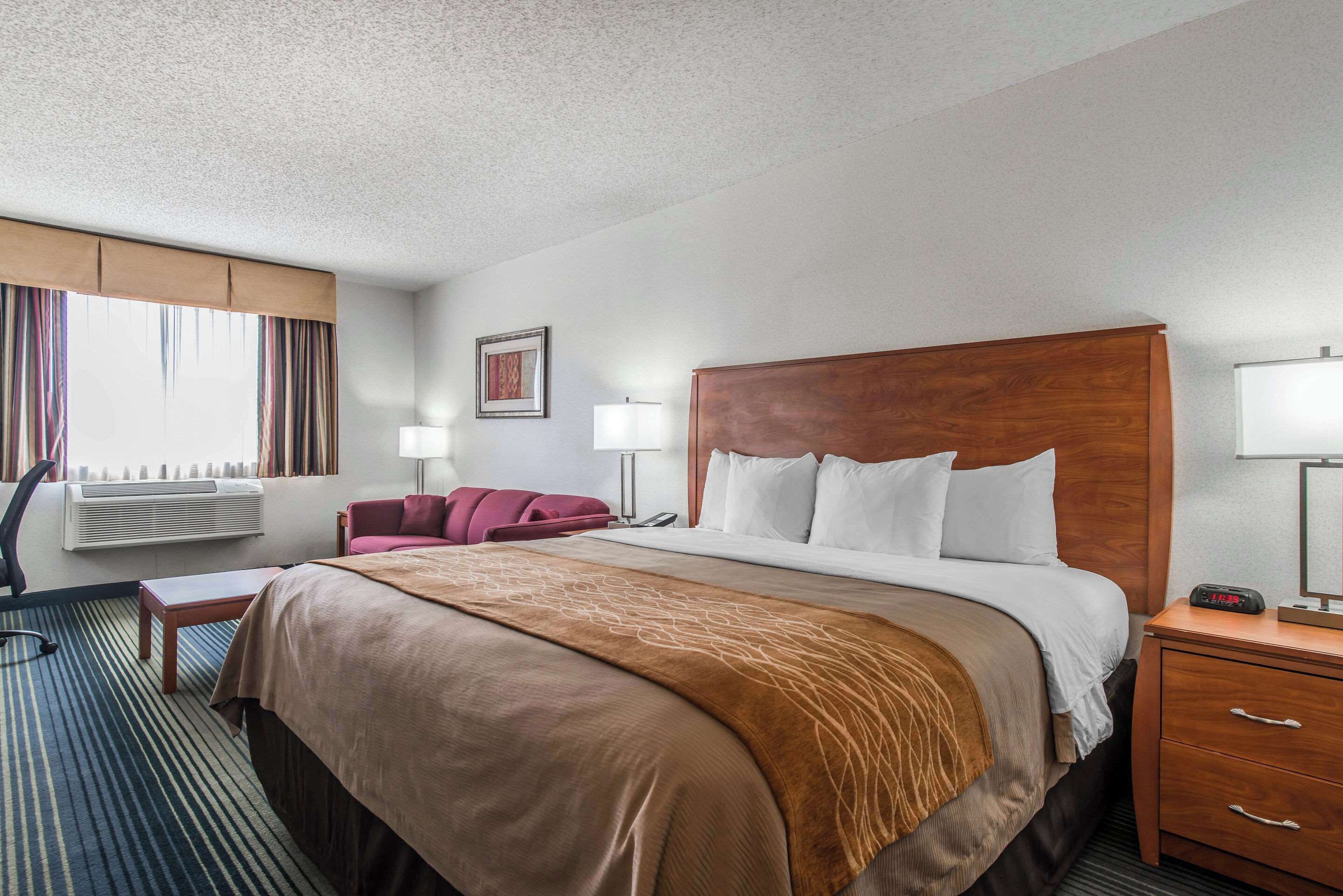 A hotel room with a large bed, a desk, a chair, a sofa, a lamp, and a clock.