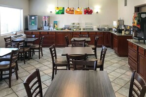 A hotel dining area with wooden tables and chairs, a coffee station, and a refrigerator.