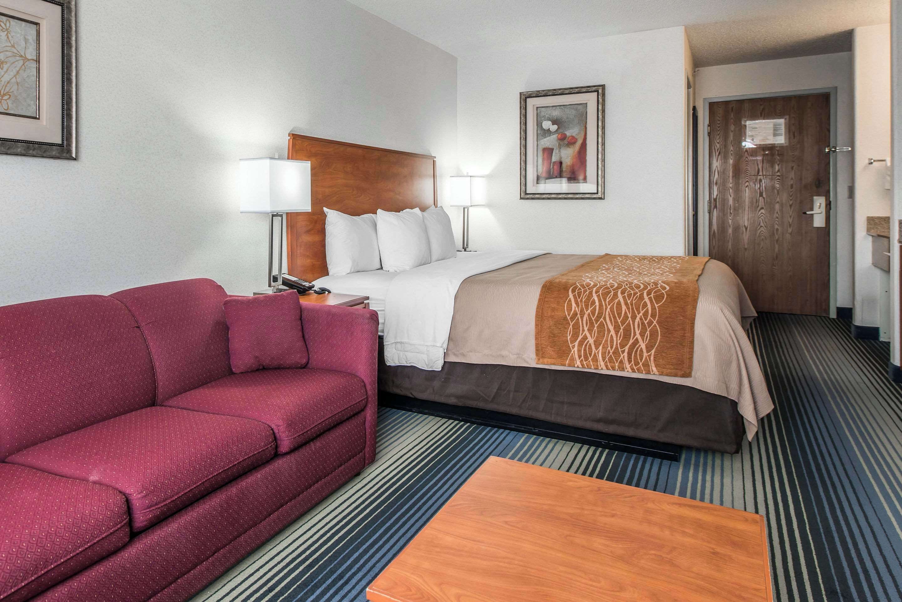 Una habitación de hotel con una cama, un sofá rojo, una mesa de centro de madera y dos lámparas fijadas a la pared.