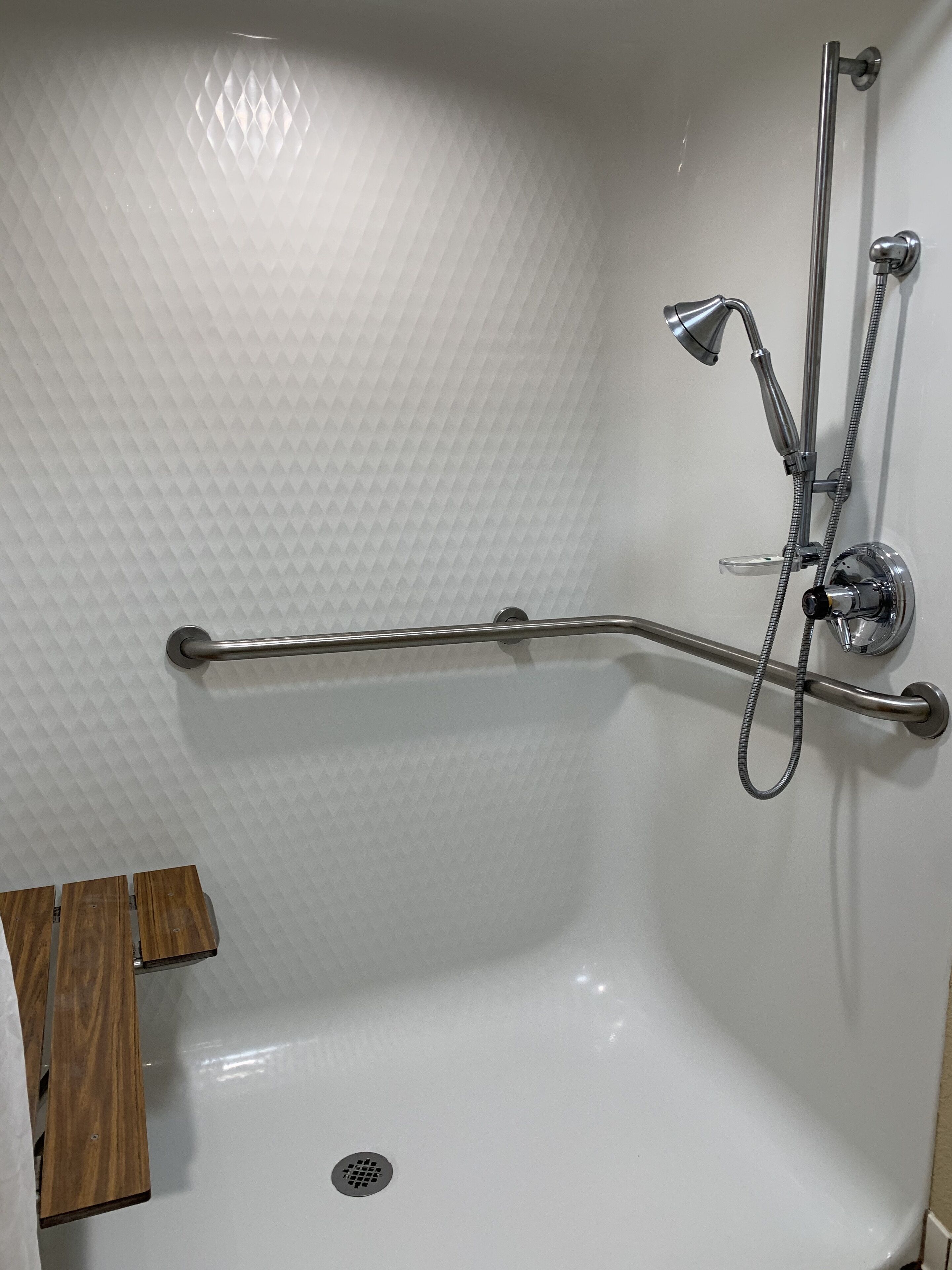 Un baño con pared blanca texturizada, ducha con cabezal de ducha manual y barra de apoyo.