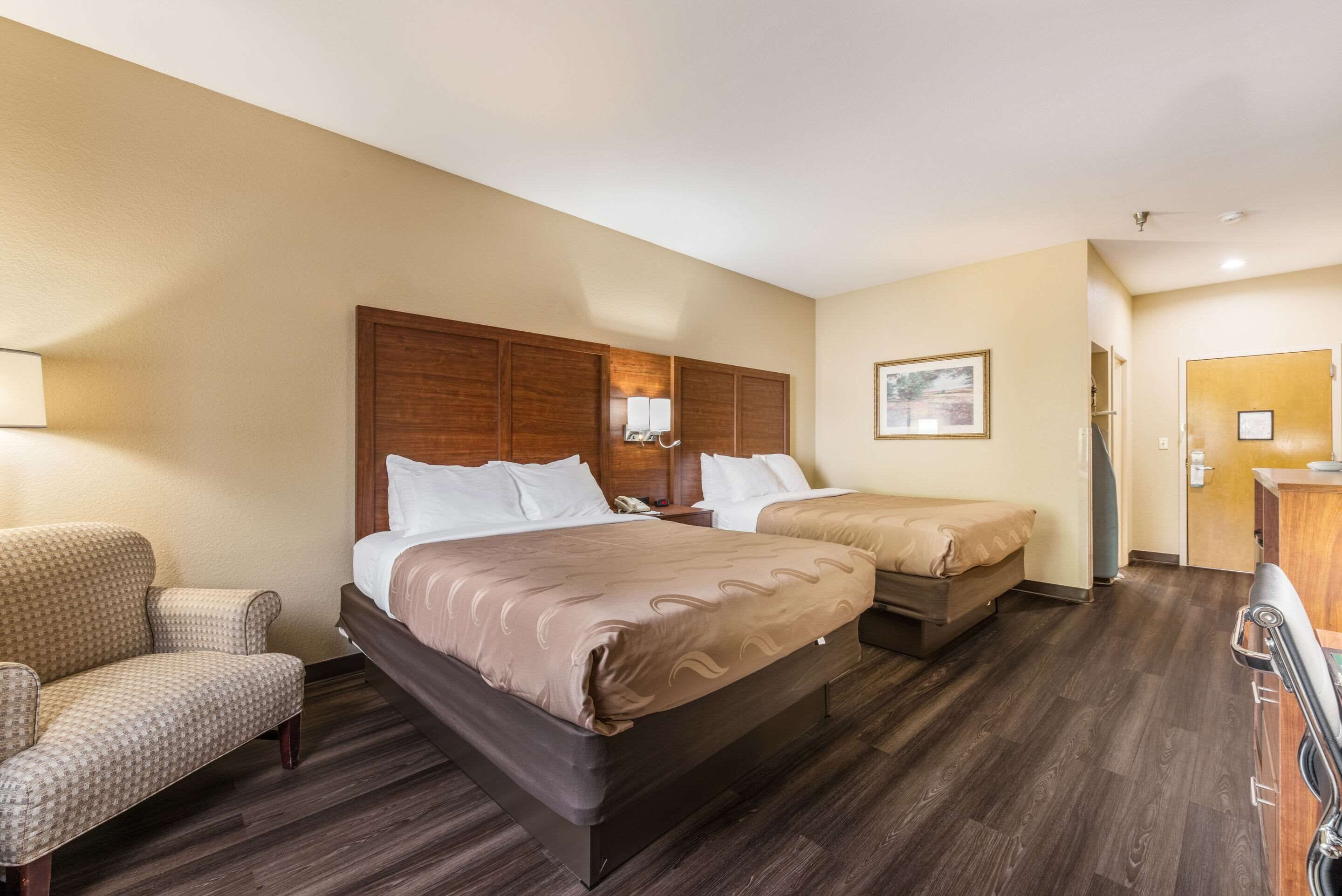 A hotel room with two beds, a chair, a desk, and a wooden headboard.
