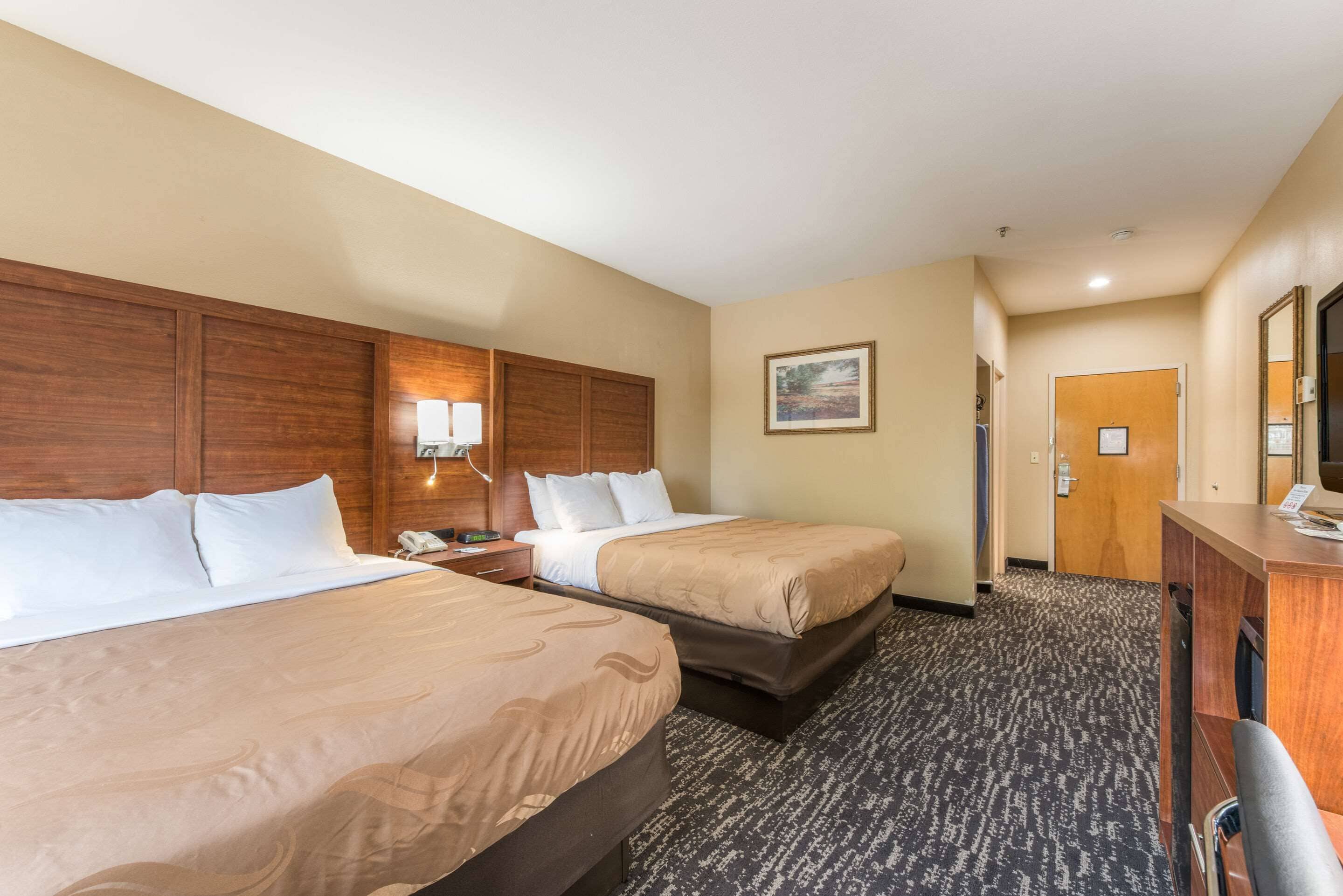 Quarto de hotel com duas camas, uma escrivaninha, uma cadeira, um abajur, um telefone e uma porta.