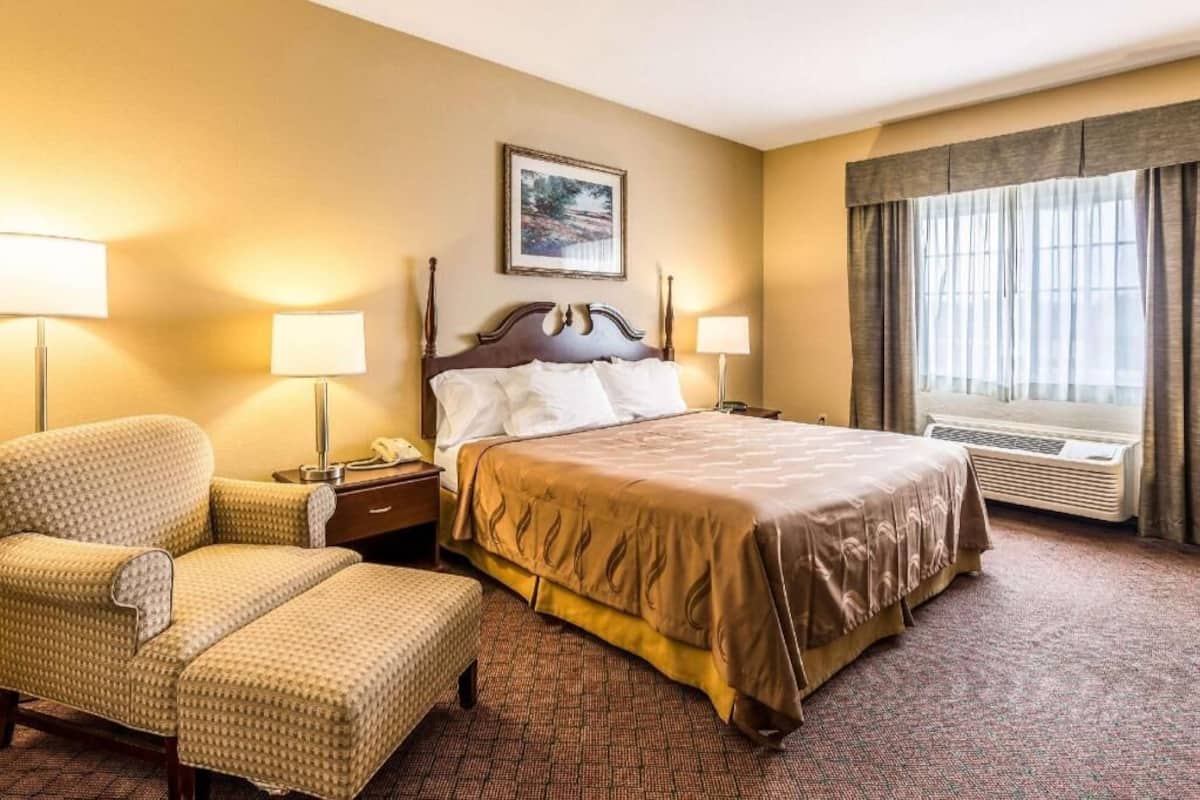 Una habitación de hotel con una cama grande, dos lámparas de noche, una silla, un escritorio y una ventana con cortinas.