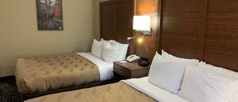 Una habitación de hotel con dos camas, una mesita de noche con teléfono y despertador, un cuadro en la pared y un cabecero de madera.
