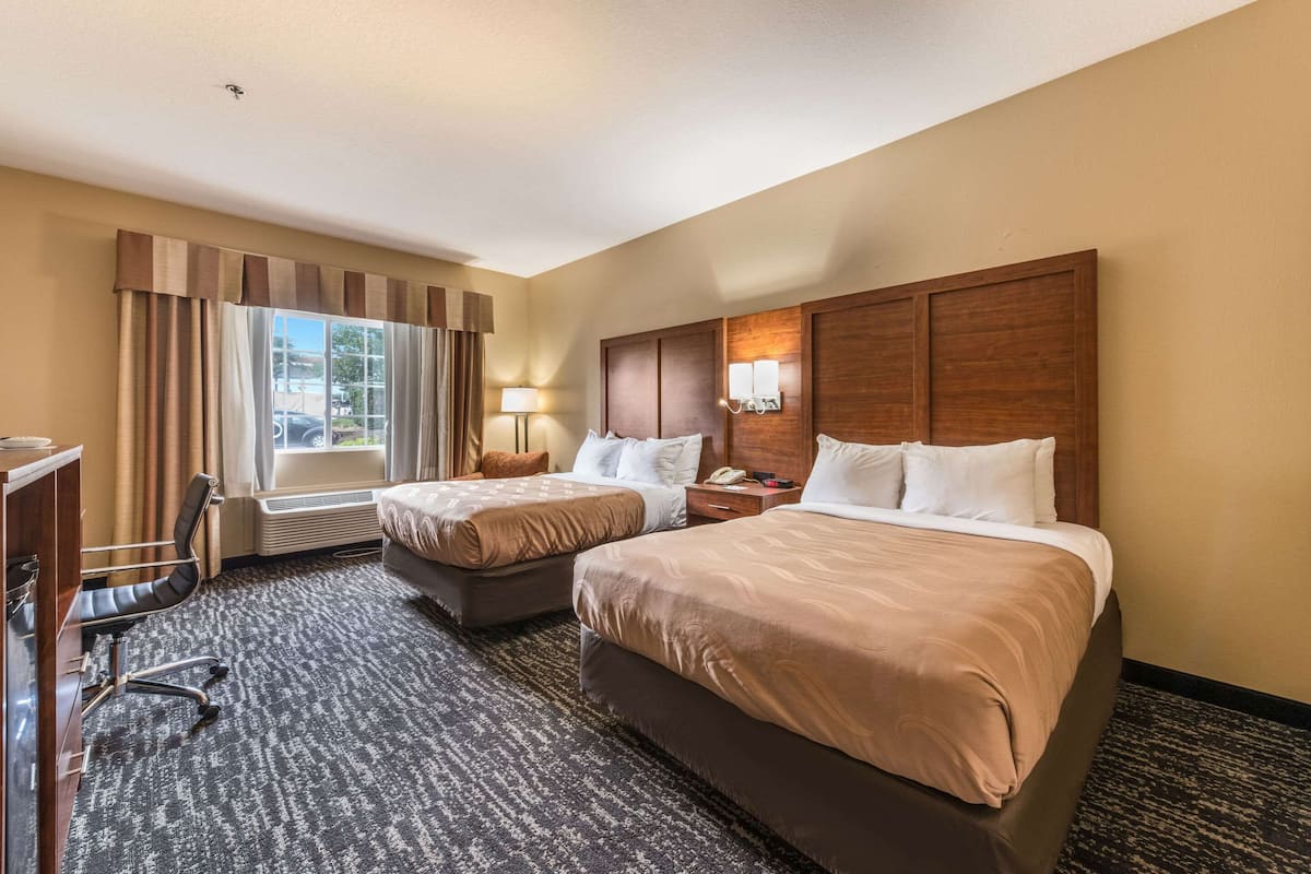Habitación de hotel con dos camas, un escritorio, una silla, una ventana con cortinas y una lámpara de pared.