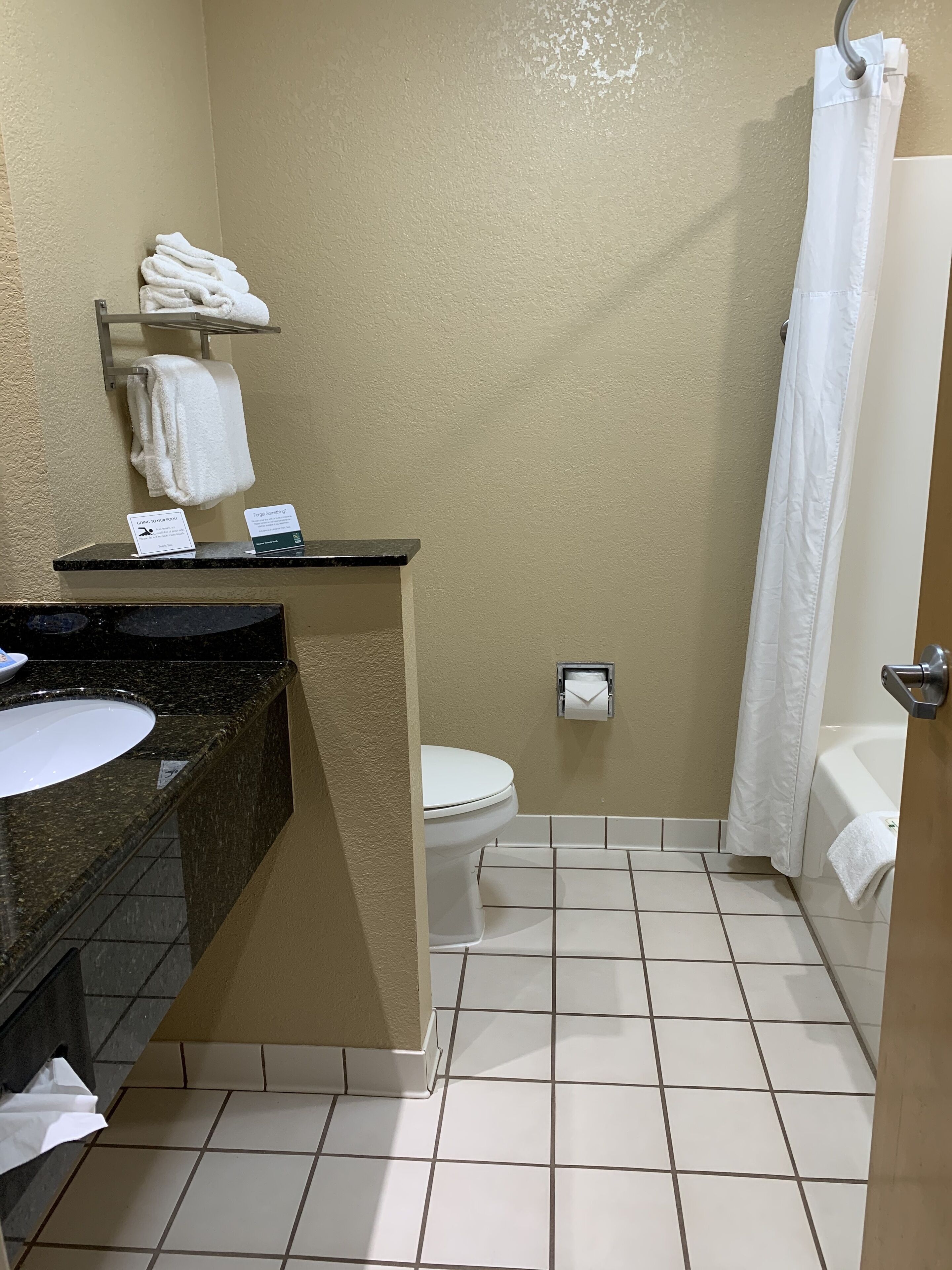 A bathroom with a sink, toilet, and shower curtain. There are towels on a shelf and a sign on the counter.