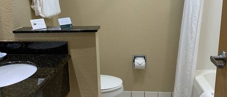 A bathroom with a sink, toilet, and shower curtain. There are towels on a shelf and a sign on the counter.