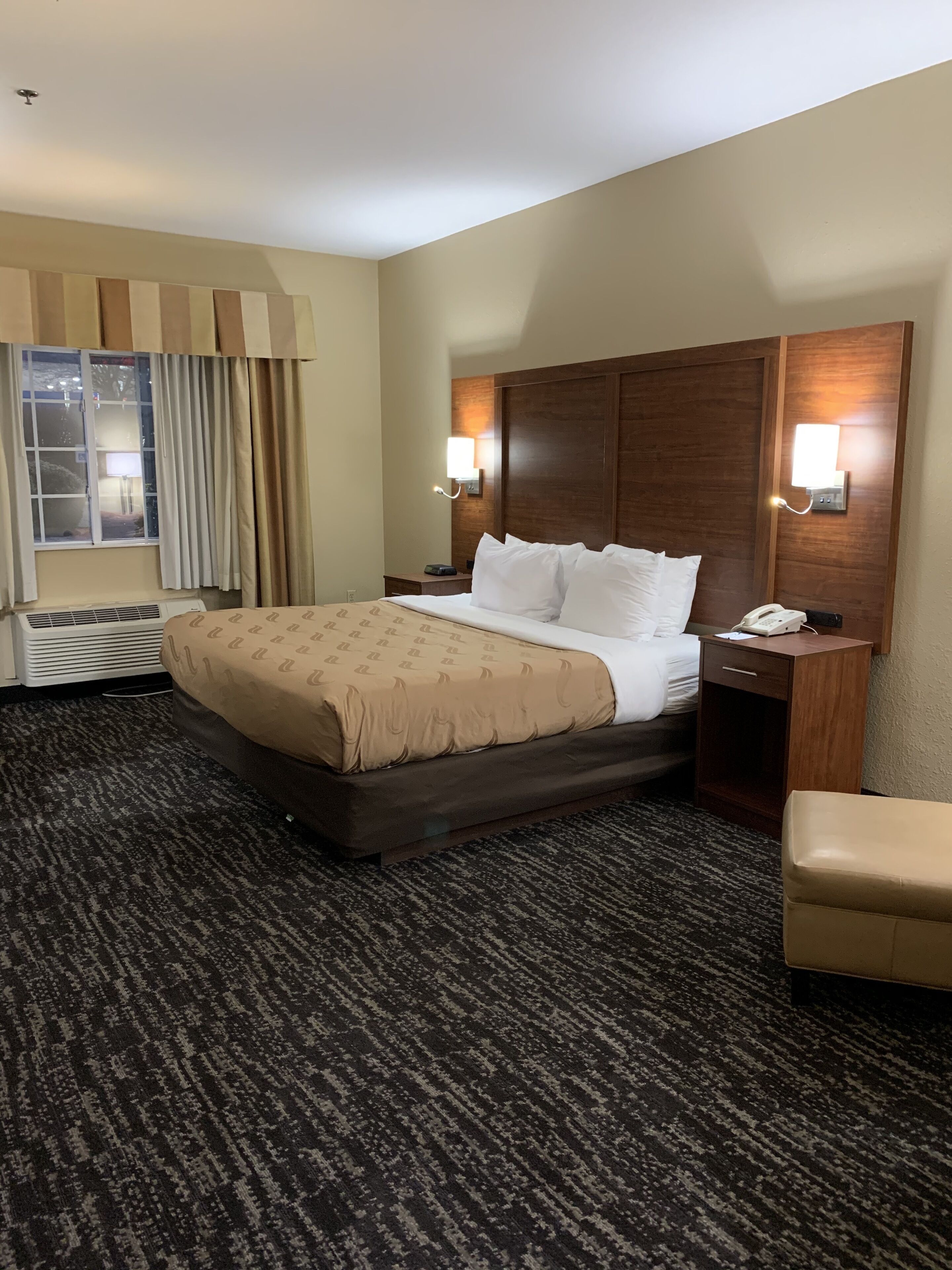 A hotel room with a large bed, two bedside tables, a chair, a window with curtains, and a wall-mounted phone.