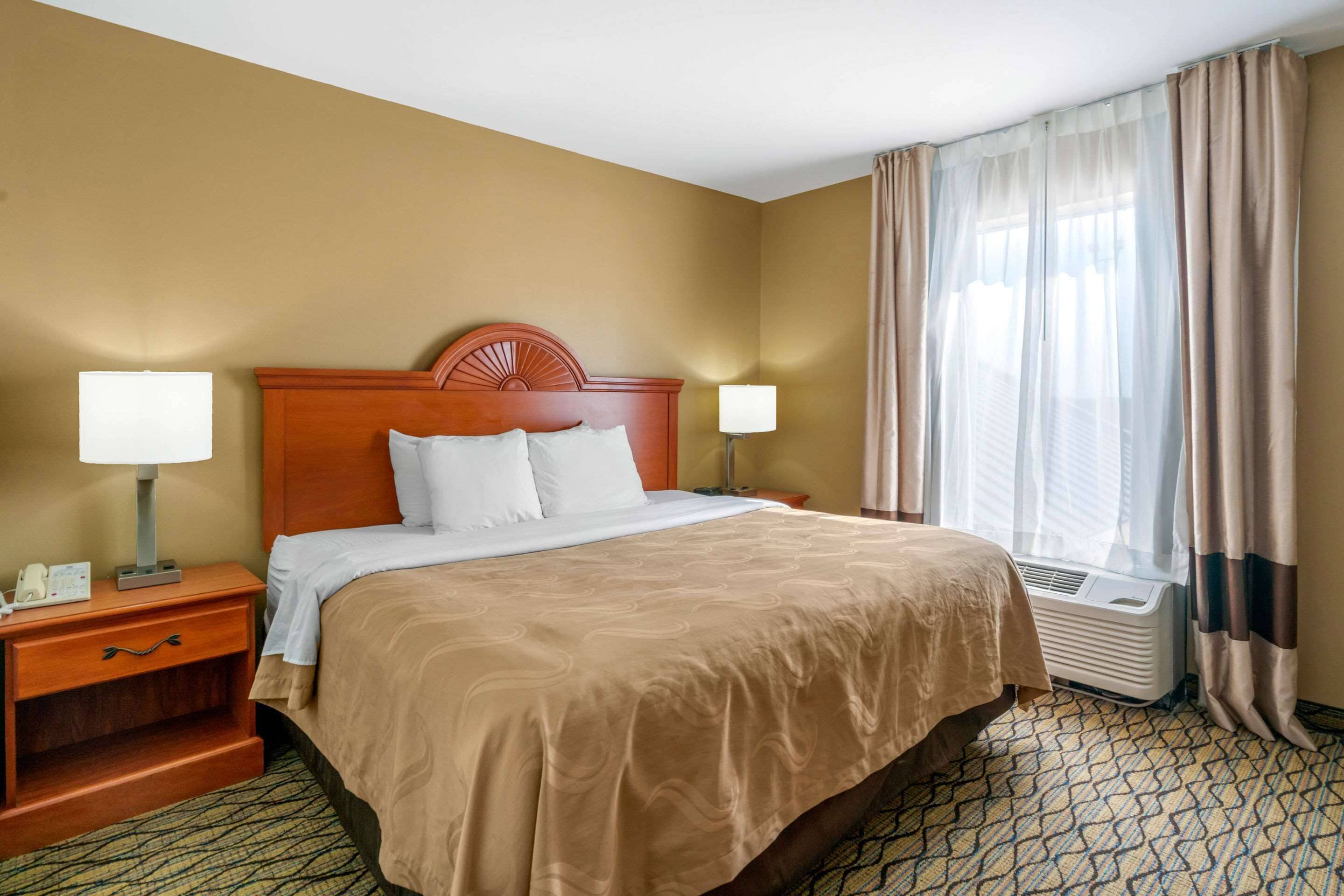 Hotelkamer met een groot bed, twee bedlampen, een nachtkastje, een raam met gordijnen en een wandairconditioner.