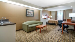 A hotel room with a kitchenette, a green sofa, a bed with a beige bedspread, a desk with a lamp, and a patterned carpet. - Quality Inn & Suites Jefferson City (Jefferson City)