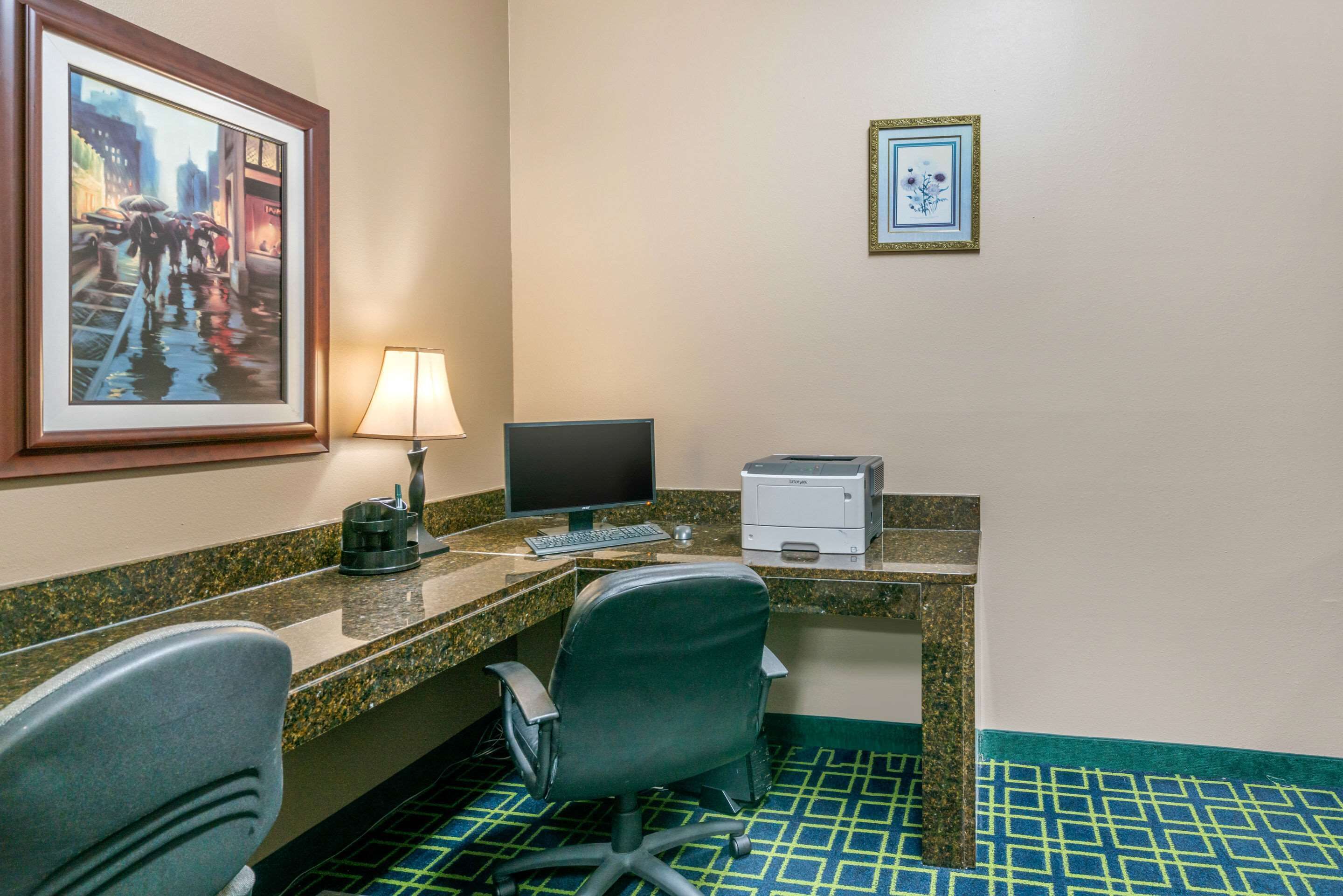 Habitación de hotel con escritorio, dos sillas, computadora, impresora, lámpara y obras de arte enmarcadas en la pared.