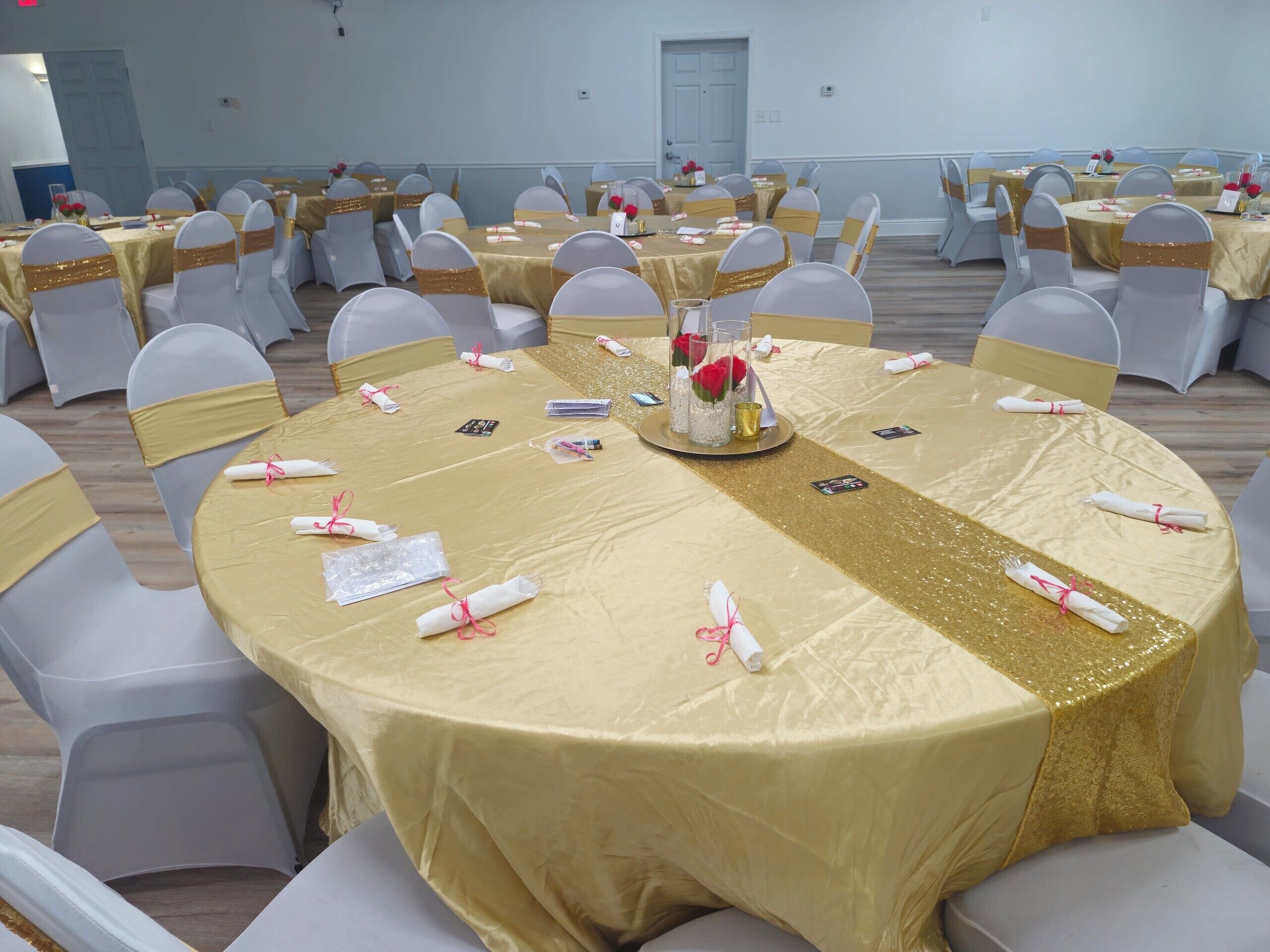 A banquet hall with round tables covered in gold tablecloths, white chairs with gold sashes, and centerpieces with red roses.