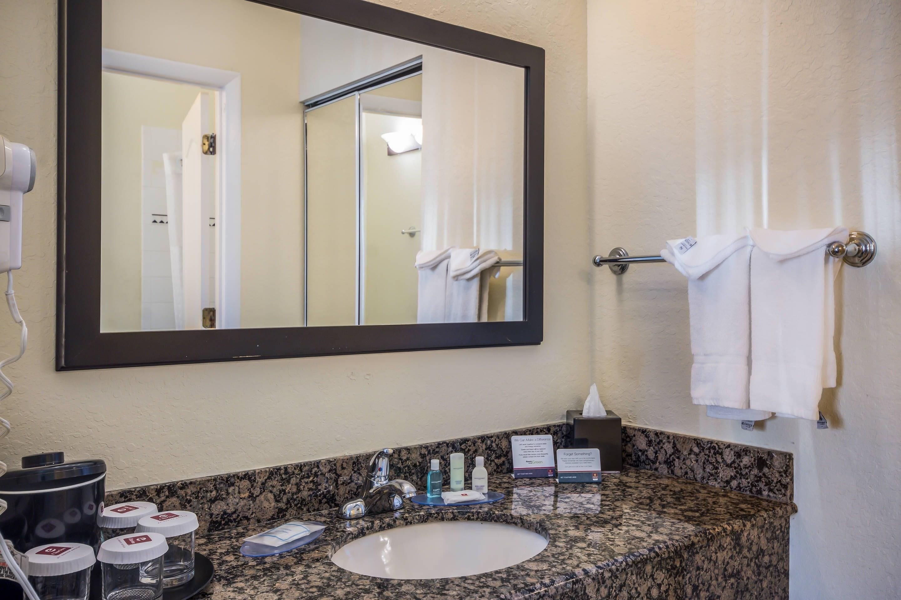 Une salle de bain d’hôtel avec un lavabo doté d’un plan de travail en granit, un grand miroir, un porte-serviettes et un panneau indiquant la présence d’un distributeur de savon sans contact.