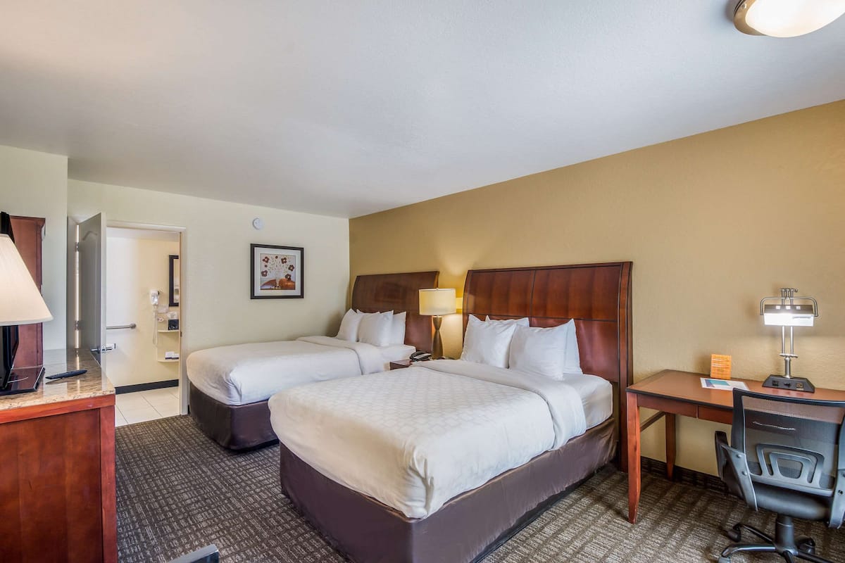 A hotel room with two beds, a desk, a chair, a lamp, and a clock on the wall.