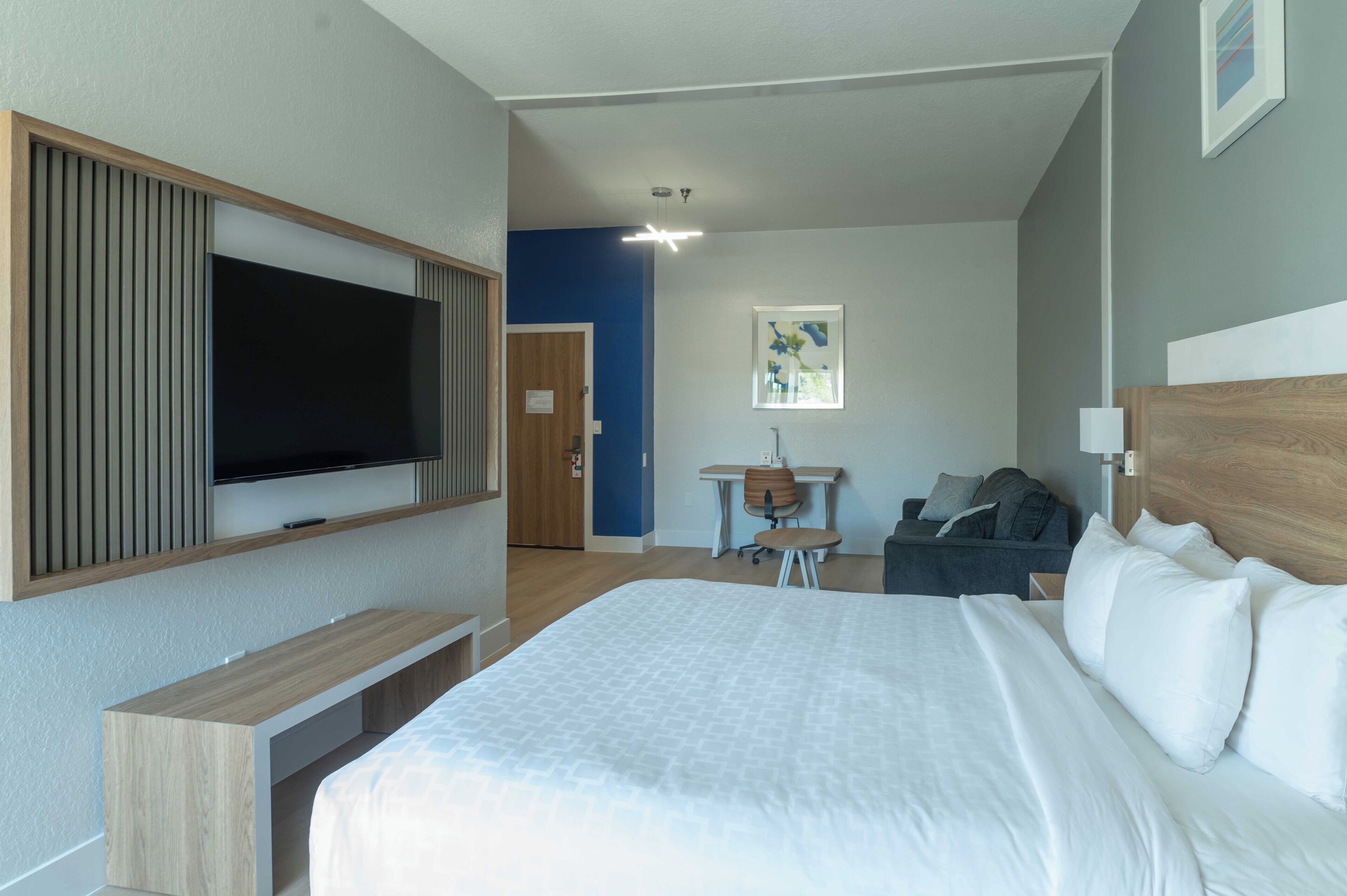 Een moderne hotelkamer met een groot bed, een aan de muur bevestigde televisie, een bureau met stoel en een kleine tafel met stoel.