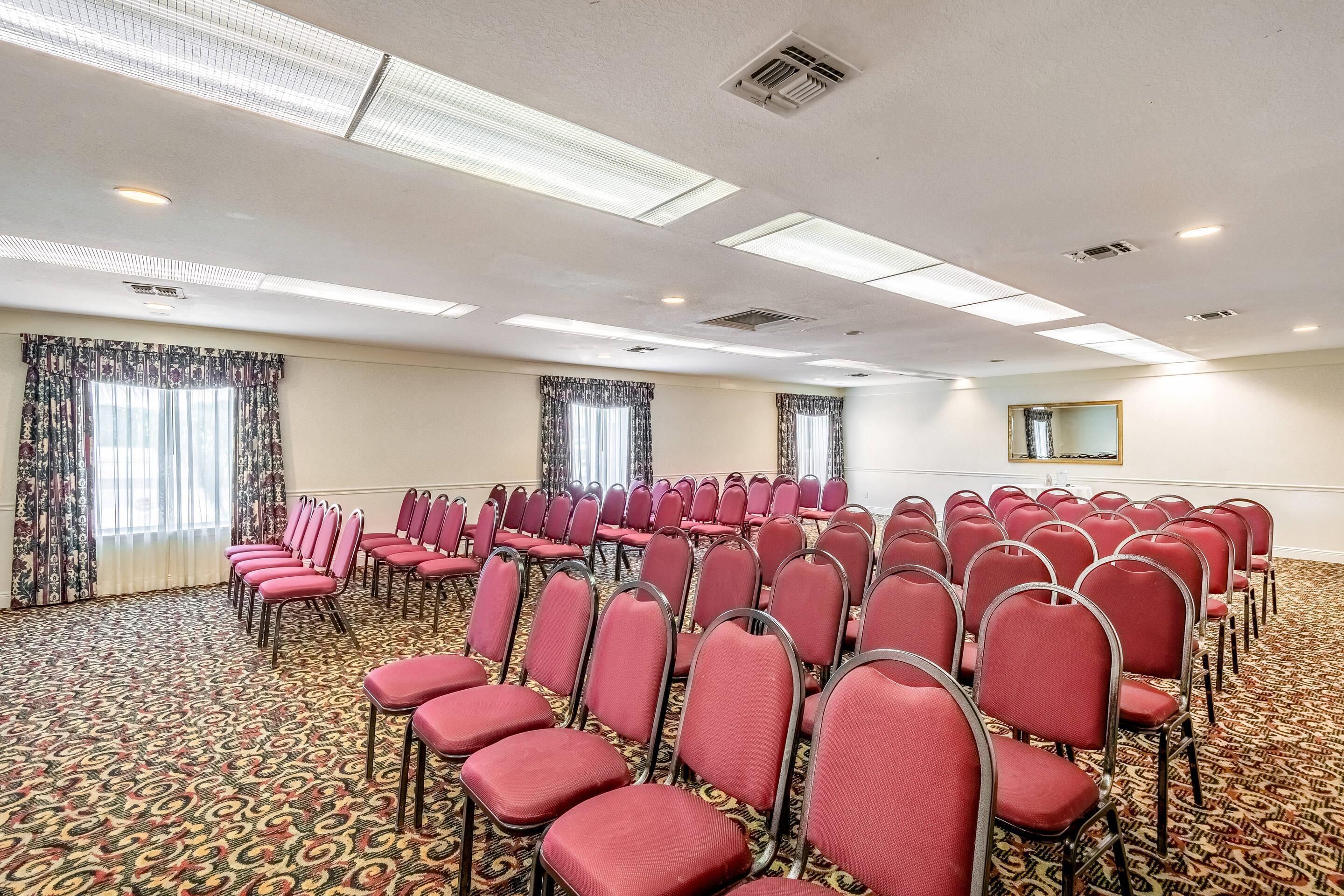 Un salón de conferencias con hileras de sillas rojas, alfombra estampada, luces de techo y ventanas con cortinas.