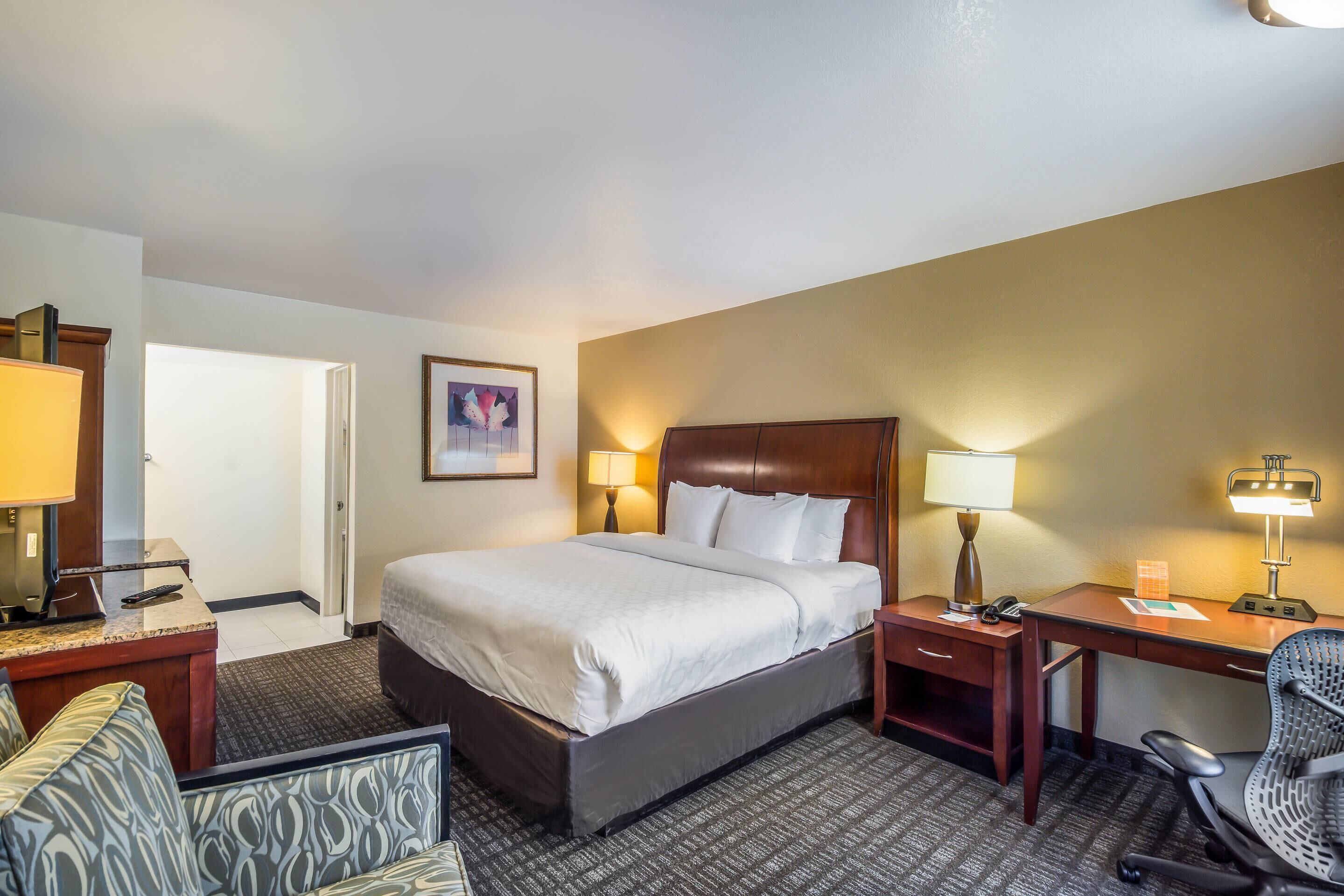 A hotel room with a bed, desk, chair, and a small table with a lamp.
