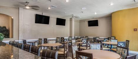 Comedor de hotel con mesas redondas, sillas acolchadas, ventiladores de techo, iluminación empotrada y televisores montados.