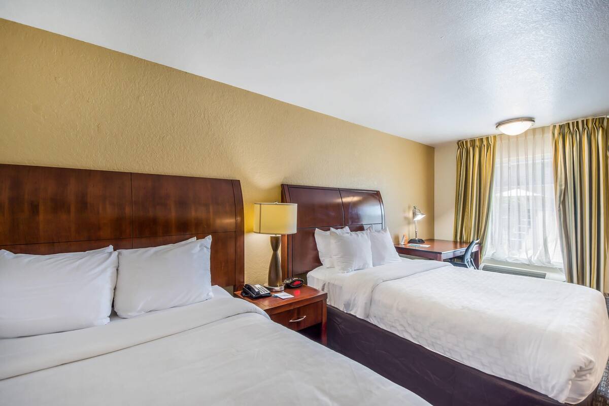 A hotel room with two beds, wooden headboards, a bedside table with a lamp, a phone, and a window with curtains.