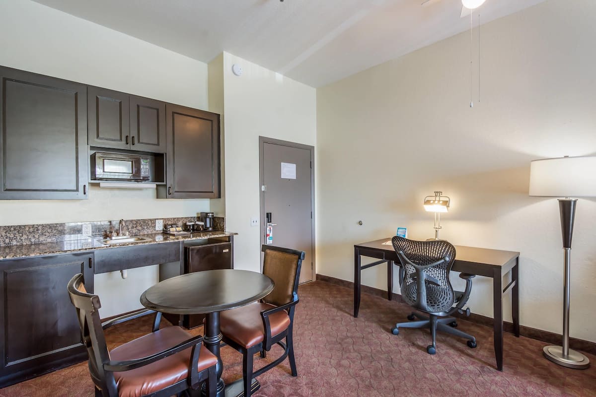 A compact kitchen with a microwave, a dining table with chairs, a desk with a chair, and a floor lamp.