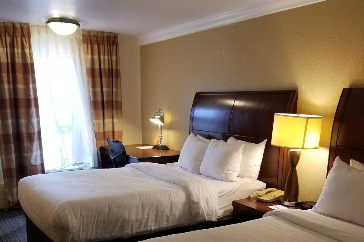 A hotel room with two beds, a desk, a chair, a lamp, and a window with curtains.
