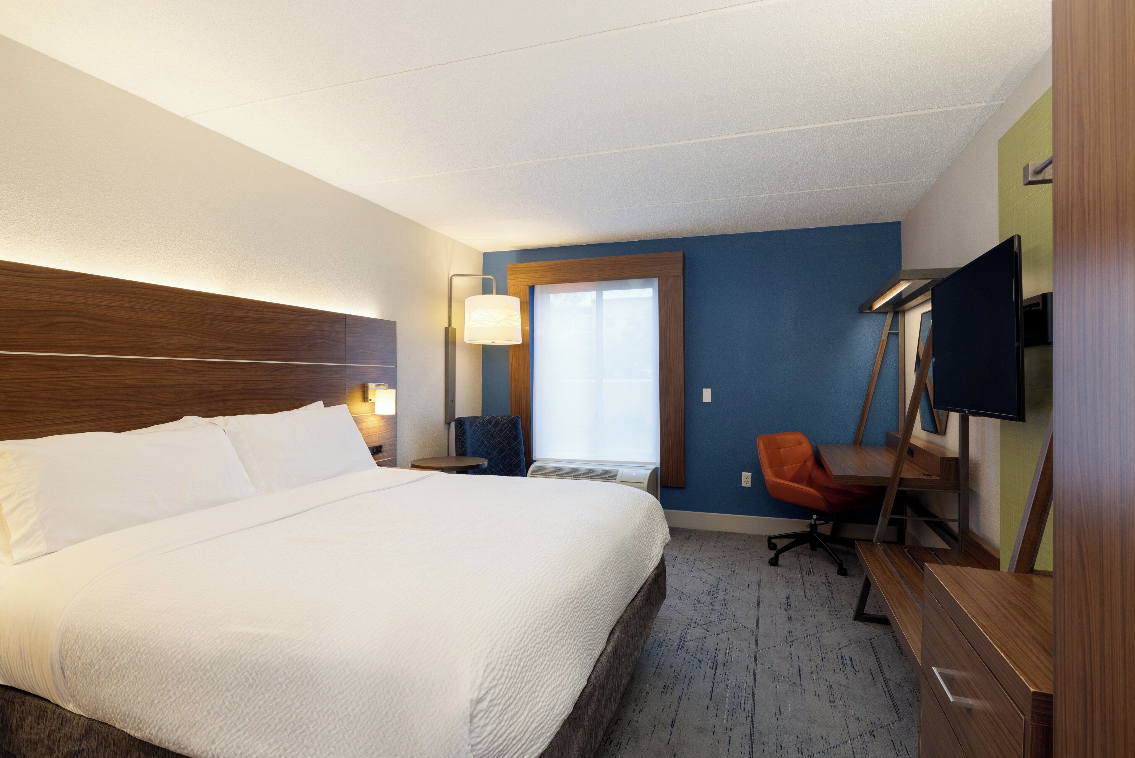 Une chambre d’hôtel avec un grand lit, un bureau avec une chaise, une télévision et une fenêtre avec des stores.